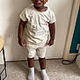 casual_clothing, child, curly_hair, emotion, expression, face, floor, footwear, furniture, human, indoor, person, room, shorts, socks, standing, table, tile_floor, wall, white_tshirt