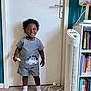 child, toddler, smiling, indoor, dinosaur_outfit, shorts, socks, bread, fan, bookshelf, books, door, floor, wall, standing, curly_hair, white_socks, happy, person, furniture