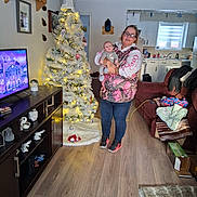 Nico is registered to the contest to win money with this photo: woman, baby, christmas_tree, living_room, television, couch, blanket, flooring, kitchen, lights, holiday, decorations, person, furniture, indoors, glasses, sneakers, toy, wood_floor, shelf