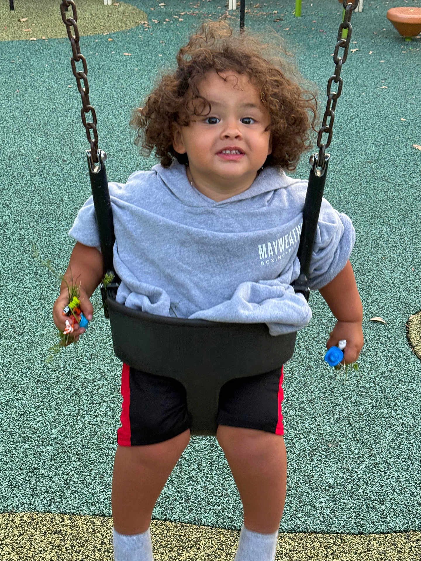 Alex joined the competition — help win amazing prizes! child, toddler, swing, playground, curly_hair, hoodie, shorts, toys, grass, outdoor, play, fun, smiling, person, recreation, seat, chains, ground, casual_clothing, happy