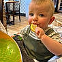 toddler, child, lemon, eating, high_chair, plush, green_overall, striped_shirt, blue_eyes, indoor, wooden_chair, table, dining_area, curious, face, person, baby, cushion, cute, food