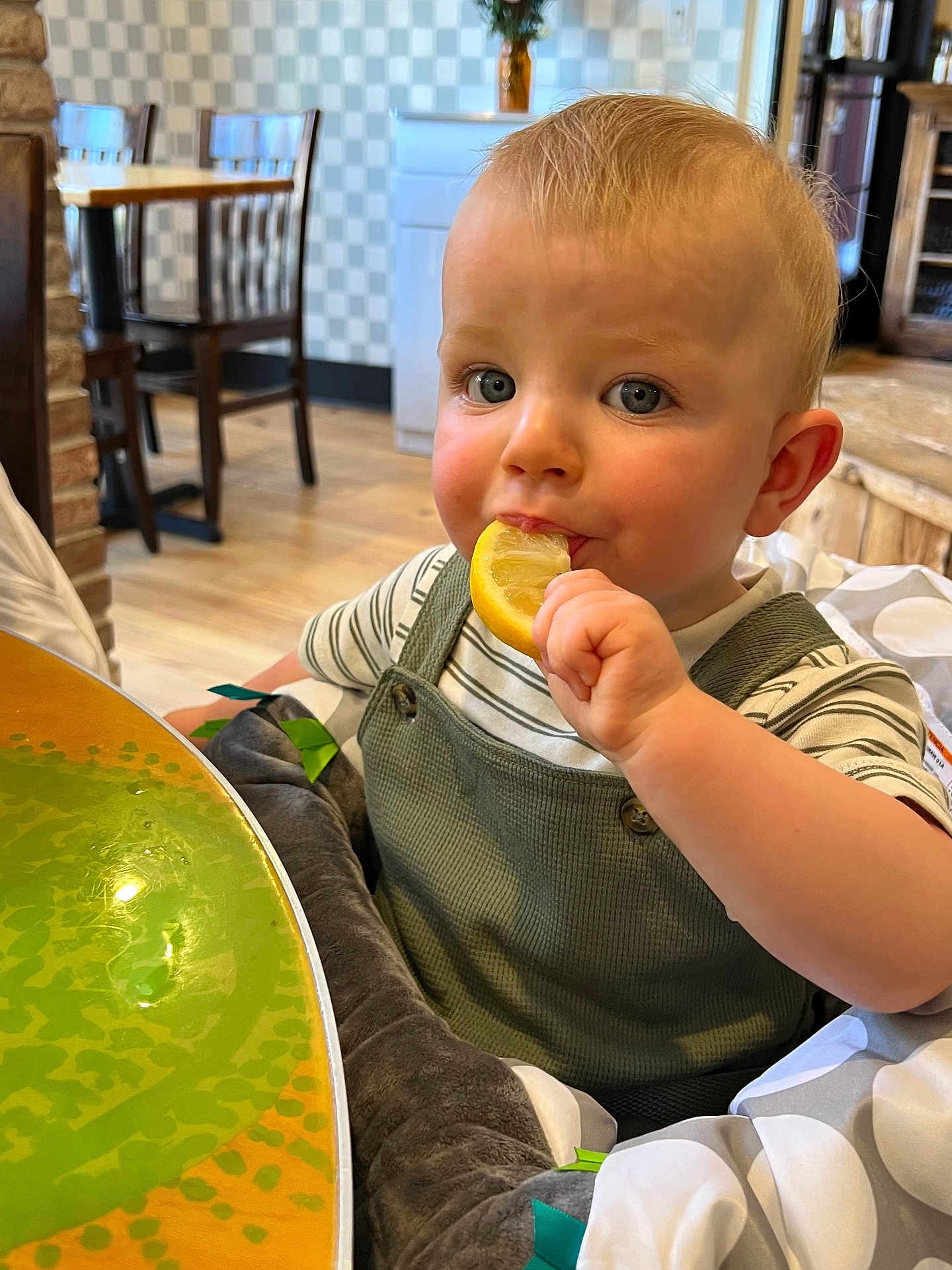 Theodore is registered to the contest to win money with this photo: toddler, child, lemon, eating, high_chair, plush, green_overall, striped_shirt, blue_eyes, indoor, wooden_chair, table, dining_area, curious, face, person, baby, cushion, cute, food