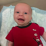 Jamezs is registered to the contest to win money with this photo: baby, infant, smiling, red_shirt, jordan_logo, closeup, face, blue_eyes, lying_down, blanket, happy, cute, child, portrait, apparel, indoor, skin, head, expression, person
