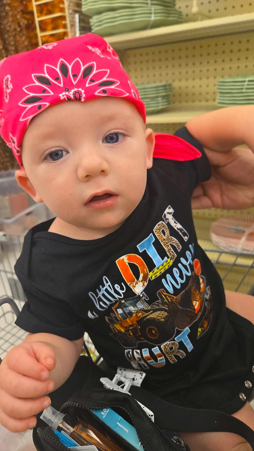 Archer is registered to the contest to win money with this photo: baby, child, bandana, shirt, construction_vehicle, shopping_cart, person, indoor, face, hand, adult_arm, bag, vape, menthol, clothing, looking_at_camera, sitting, retail, shelf, plate