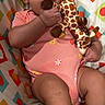baby, chewing, child, colorful, cushion, cute, expression, face, giraffe, hand, indoors, infant, legs, lying_down, onesie, pattern, pink_clothing, plush_toy, soft_toy, young_child