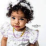 child, toddler, baby, curly_hair, headband, floral_dress, purple_lace, amber_necklace, big_eyes, eyelashes, studio_portrait, white_background, wooden_chair, close_up, expressive_face, adorable, sitting, innocence, soft_lighting, photographer_watermark