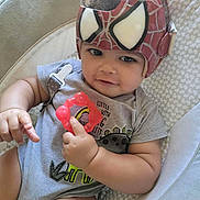Uriah joined the competition — help win amazing prizes! baby, child, helmet, spiderman, teether, gray_onesie, curly_hair, indoor, cushion, soft_bedding, cute, smiling, holding, infant, person, closeup, relaxed, toy, headgear, comfortable