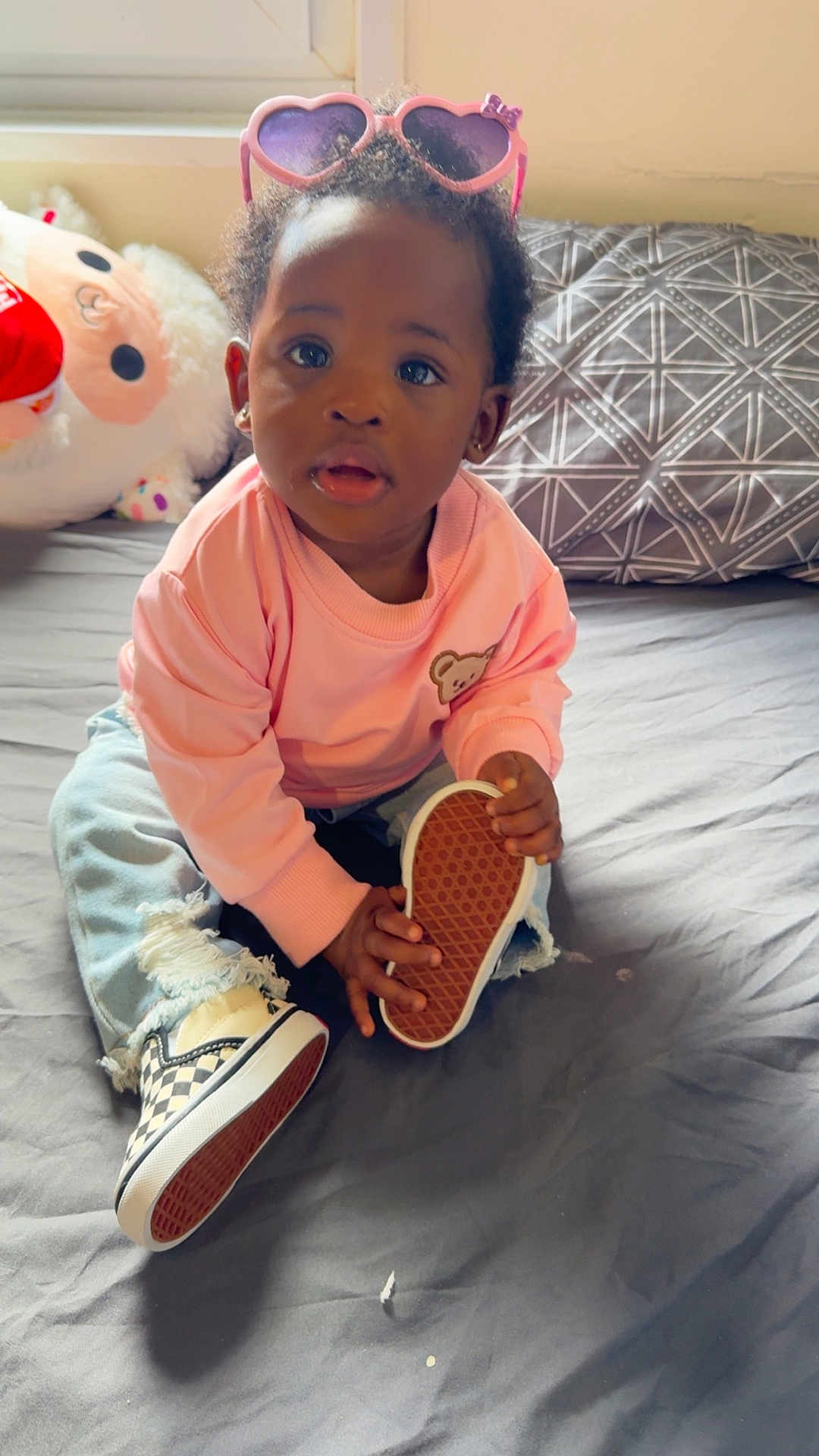 Ryla is registered to the contest to win money with this photo: toddler, child, sunglasses, pink_sweater, ripped_jeans, shoe, bed, pillow, stuffed_animal, indoor, person, casual_clothing, cute, sitting, holding, footwear, young_child, home, portrait, curly_hair