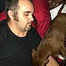 man, dog, sleeping, resting, tattoo, piercing, pillow, red, black_shirt, brown_dog, indoor, bed, closeup, relaxation, pet, animal, companion, human, face, hair