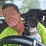 man, dog, car, steering_wheel, seat, green_shirt, black_dog, white_spots, collar, dog_tag, window, tree, road_sign, inside, portrait, close_up, pet, animal, vehicle_interior, casual