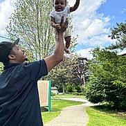 Ryan is registered to the contest to win money with this photo: baby, child, adult, man, outdoor, park, grass, trees, sky, clouds, sidewalk, smile, playing, holding, person, casual_clothing, hat, nature, daylight, joy