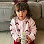 child, toddler, girl, bottle, sitting, couch, sweater, bows, hair_buns, bangs, big_eyes, portrait, indoors, floor, red_pants, white_bottle, fluffy_clothing, cute, soft_lighting, shallow_depth_of_field