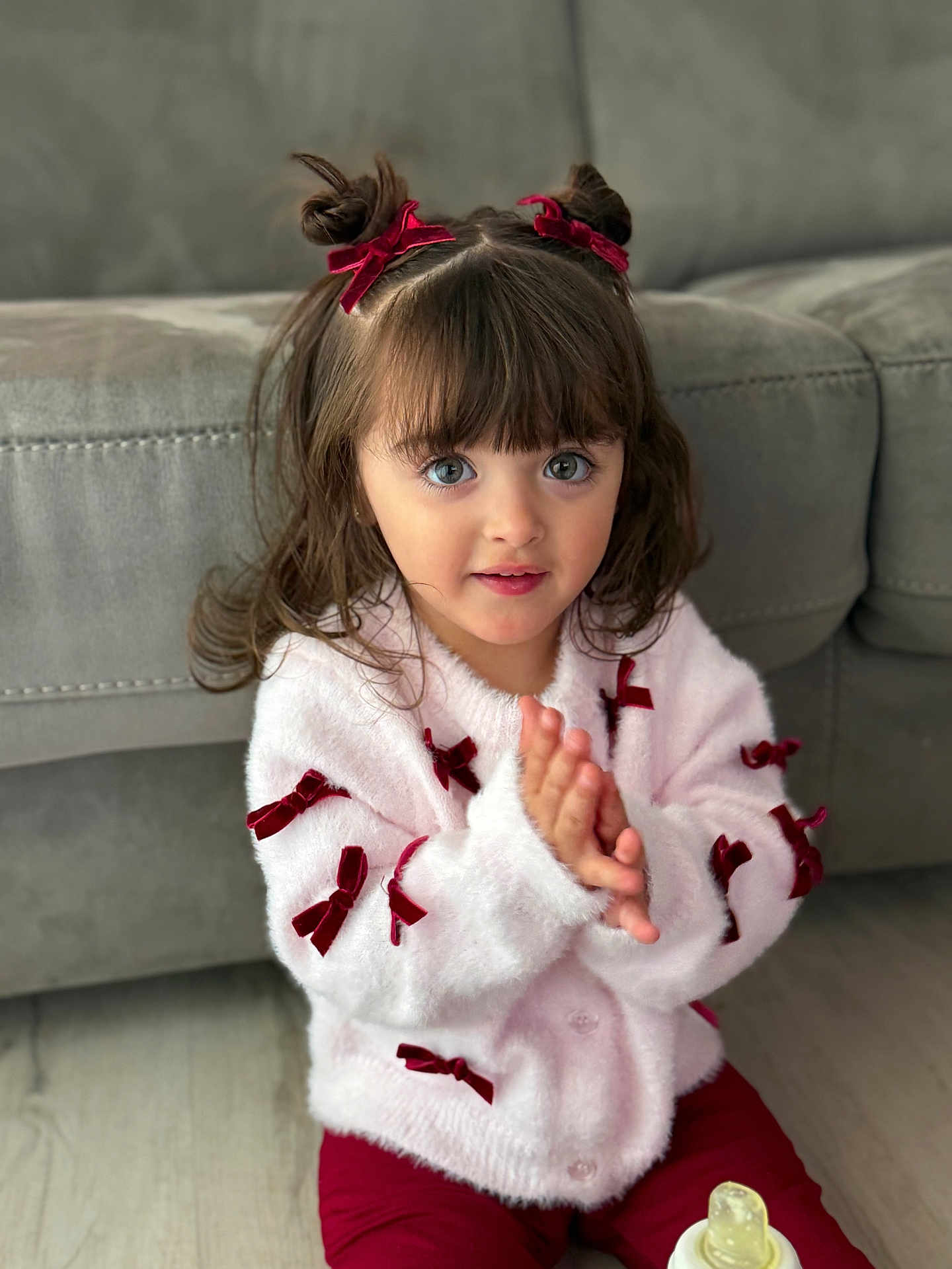 Mía Blanca joined the competition — help win amazing prizes! child, toddler, girl, bow, hair_buns, ribbon, sweater, pink_sweater, red_bows, sofa, couch, living_room, baby_bottle, sitting, hands, big_eyes, portrait, indoor, cute, floor