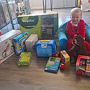 Kayden a rejoint le concours — aidez-le/la à gagner de superbes lots ! toddler, child, red_clothing, blue_chair, stuffed_animal, monkey_toy, toys, boxes, wooden_floor, window, table, furniture, indoor, happy, smiling, person, playtime, living_room, daylight, home