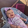 adorable, baby, baby_seat, blue_eyes, blurred_background, bow, cheeks, close_up, happy, headband, infant, onesie, portrait, print_on_clothing, purple_clothing, reclined, small_hands, smiling, soft_lighting, wooden_deck