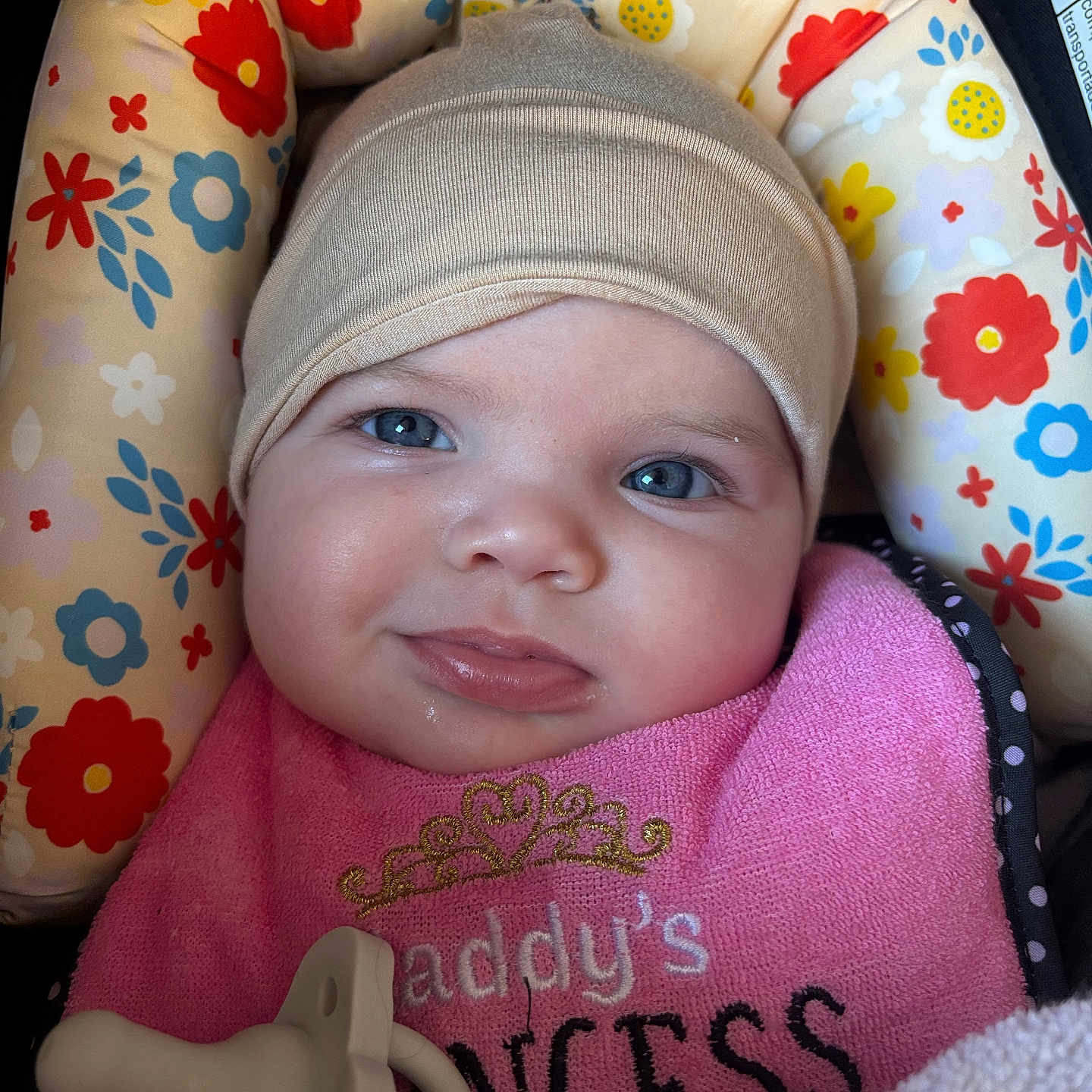 Mikiah is registered to the contest to win money with this photo: accessory, baby, beanie, bib, blue_eyes, car_seat, cheeks, child, close_up, clothing, cozy, cute, face, floral_pattern, infant, lips, pacifier, pink, portrait, smile