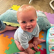 Oliver is registered to the contest to win money with this photo: baby, child, smiling, indoor, playmat, toy, plush_toy, foot, hand, colorful, floor, crawling, person, cute, infant, clothing, musical_toy, happy, blue_eyes, playroom