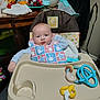 Carter is registered to the contest to win money with this photo: baby, high_chair, bib, toy, table, bottle, plastic_container, face, indoor, clothing, feeding, household, furniture, baby_toy, child, person, blue, yellow, messy_face, cosco