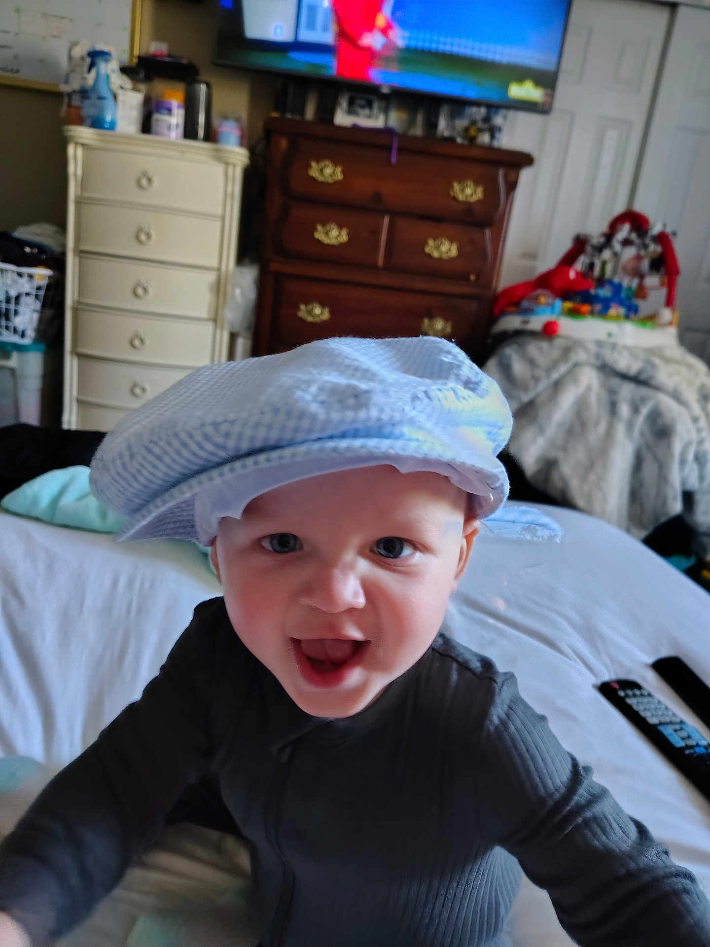 Carter joined the competition — help win amazing prizes! baby, child, hat, bed, furniture, television, remote_control, toy, drawer, blanket, room, indoor, clothing, face, expression, person, smile, happy, home, living_room