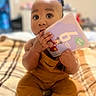 baby, child, infant, sitting, bed, blanket, book, alphabet_book, colorful, yellow_clothing, indoors, home, portrait, face, eyes, hands, chewing, curious, cozy, smiling