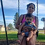 woman, baby, swing, pacifier, outdoor, park, basketball_hoop, fence, grass, tree, cloud, sky, person, holding, tattoo, sunlight, playing, child, summer, casual_clothing
