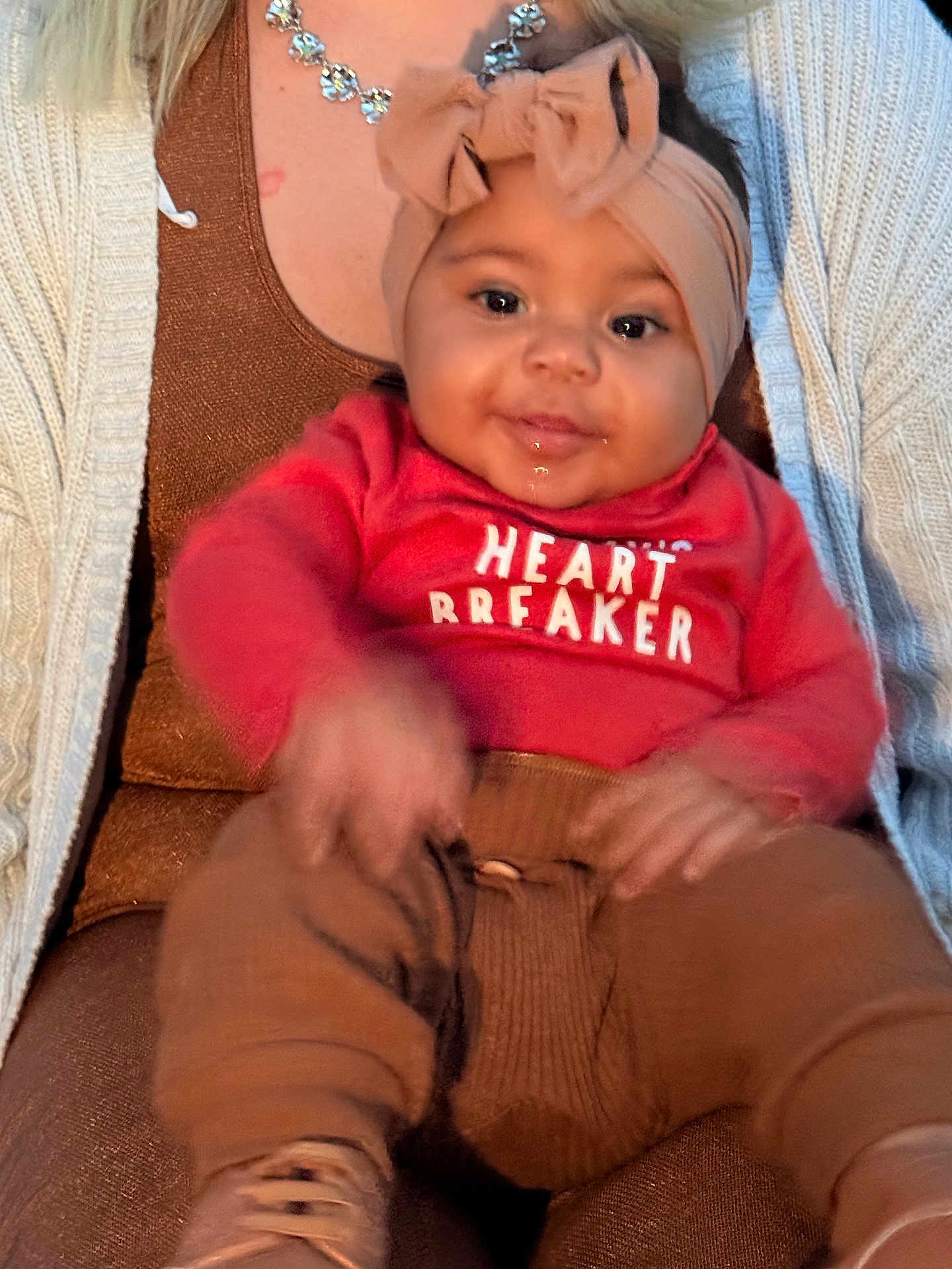 Malaina is registered to the contest to win money with this photo: baby, child, smile, headband, bow, drool, red_shirt, heart_breaker, pants, shoes, sitting, person, adult, cardigan, necklace, brown_clothing, indoor, closeup, happy, cute