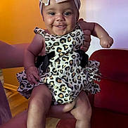Malaina is registered to the contest to win money with this photo: baby, child, smiling, leopard_print, headband, bow, flower_booties, person, indoor, red_chair, hand, clothing, cute, happy, portrait, infant, funny_expression, socks, floor, background