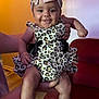 baby, child, smiling, leopard_print, headband, bow, flower_booties, person, indoor, red_chair, hand, clothing, cute, happy, portrait, infant, funny_expression, socks, floor, background