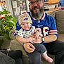 Jacen joined the competition — help win amazing prizes! man, baby, child, smiling, glasses, beard, cap, hat, sports_jersey, couch, living_room, plant, kitchen, refrigerator, carpet, casual_clothing, indoor, family, happy, seated
