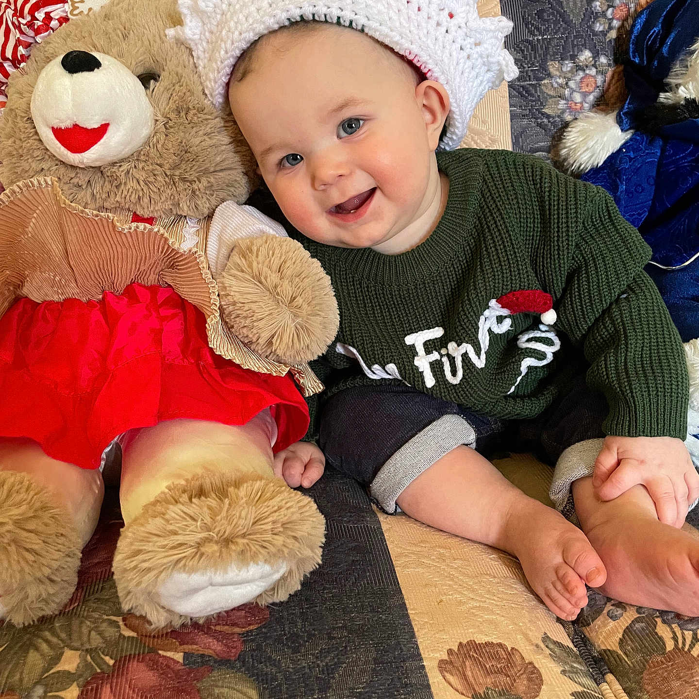 Jacen is registered to the contest to win money with this photo: baby, child, couch, cozy, crown, cute, festive, floral_pattern, footwear_free, green_sweater, happy, holiday, indoors, knitted_hat, plush_toy, portrait, red_skirt, sitting, smiling, teddy_bear