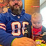 Jacen is registered to the contest to win money with this photo: adult, baby, beard, blue_jersey, child, clothing, face, fidget_spinner, glasses, hat, indoor, man, person, restaurant, table, toy, wood_paneling, window, young_child, hand