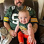man, baby, smile, green_bay_packers, jersey, glasses, beard, orange_pants, tank_top, leather_chair, blanket, indoor, happy, holding, family, portrait, casual, relaxed, person, home