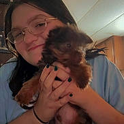 Teddy is registered to the contest to win money with this photo: person, glasses, puppy, dog, pet, holding, smile, hands, painted_nails, long_hair, tshirt, indoor, ceiling_tiles, wooden_cabinet, portrait, closeup, brown_fur, cute, home, blurred_motion