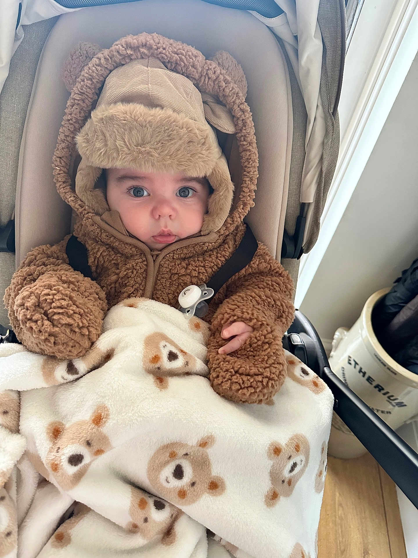 Harrison joined the competition — help win amazing prizes! baby, child, stroller, blanket, bear_pattern, brown_clothing, fuzzy_jacket, hat, pacifier, indoor, floor, wooden_floor, person, cute, warm_clothing, face, eyes, hand, infant, cozy