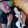 man, cat, sleeping, tattoo, couch, indoor, relaxation, black_shirt, white_cat, black_cat, neon_collar, resting, hand_on_chest, mustard_sofa, tattooed_arm, beard, closed_eyes, comfort, nap, pet