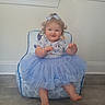 Violet is registered to the contest to win money with this photo: baby, child, toddler, portrait, sitting, smiling, happy, blue_tutu, hair_bow, dress, chair, indoor, home, window, wall, floor, cartoon_pattern, barefoot, playful, face