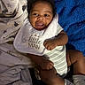 baby, infant, child, smiling, portrait, lying_down, bib, onesie, blanket, cozy, indoor, bed, blue_blanket, striped_clothing, socks, dark_skin, happy, face, baby_clothing, photograph