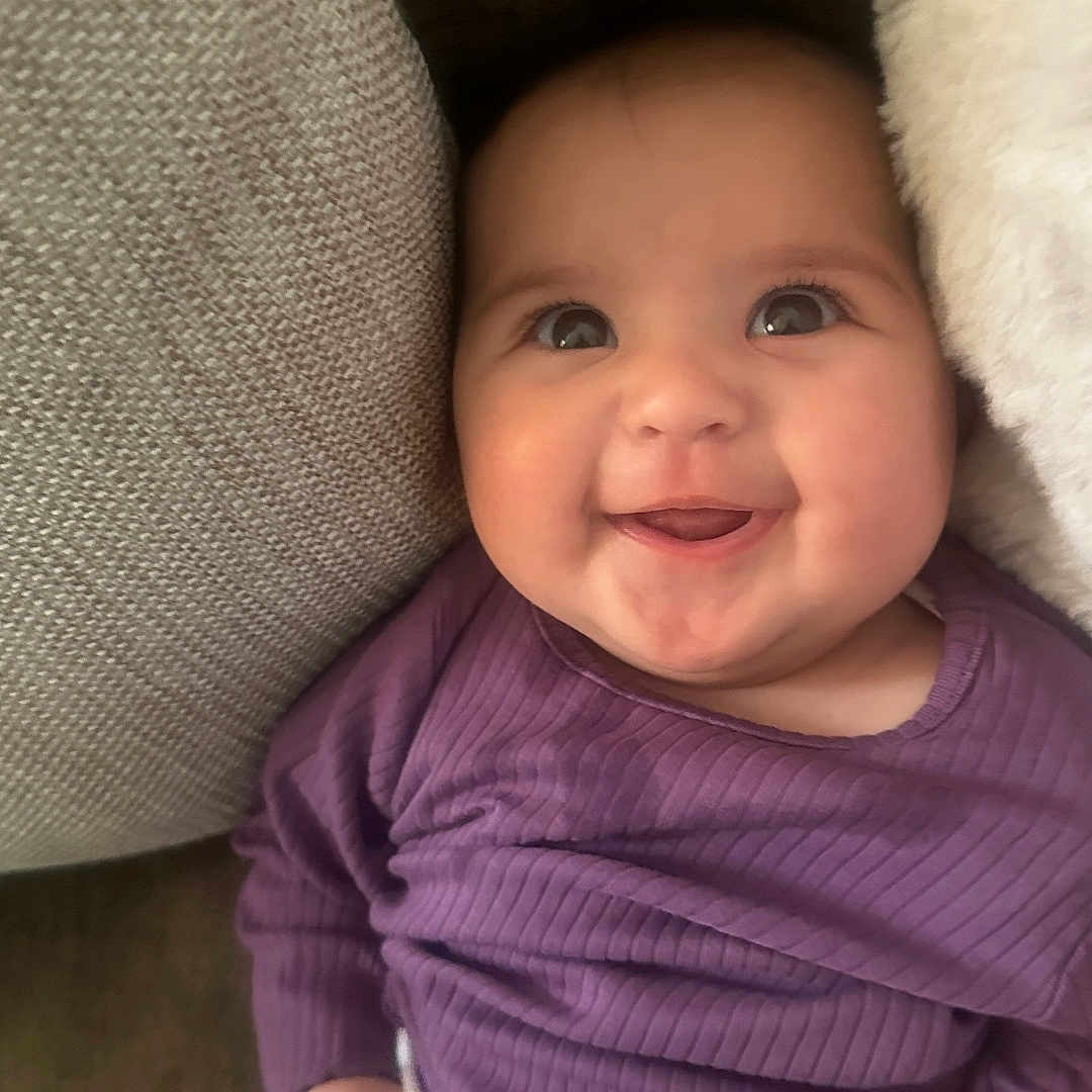 Noora is registered to the contest to win money with this photo: baby, child, closeup, comfort, couch, cozy, cushion, cute, face, happy, indoors, infant, person, portrait, purple_clothing, seated, smiling, soft_texture, warm, young_child