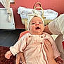 baby, infant, child, person, pink_clothing, headband, hand, indoor, wicker_bassinet, blanket, adult_hand, carpet, wall, curtain, window, chair, soft_lighting, portrait, cute, happy