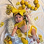 baby, infant, yellow_bow, lemon, lemon_slice, white_cushion, yellow_clothing, diaper, flower_decoration, fabric_background, cute, portrait, smiling, indoors, soft_texture, child, fruit, colorful, playful, resting