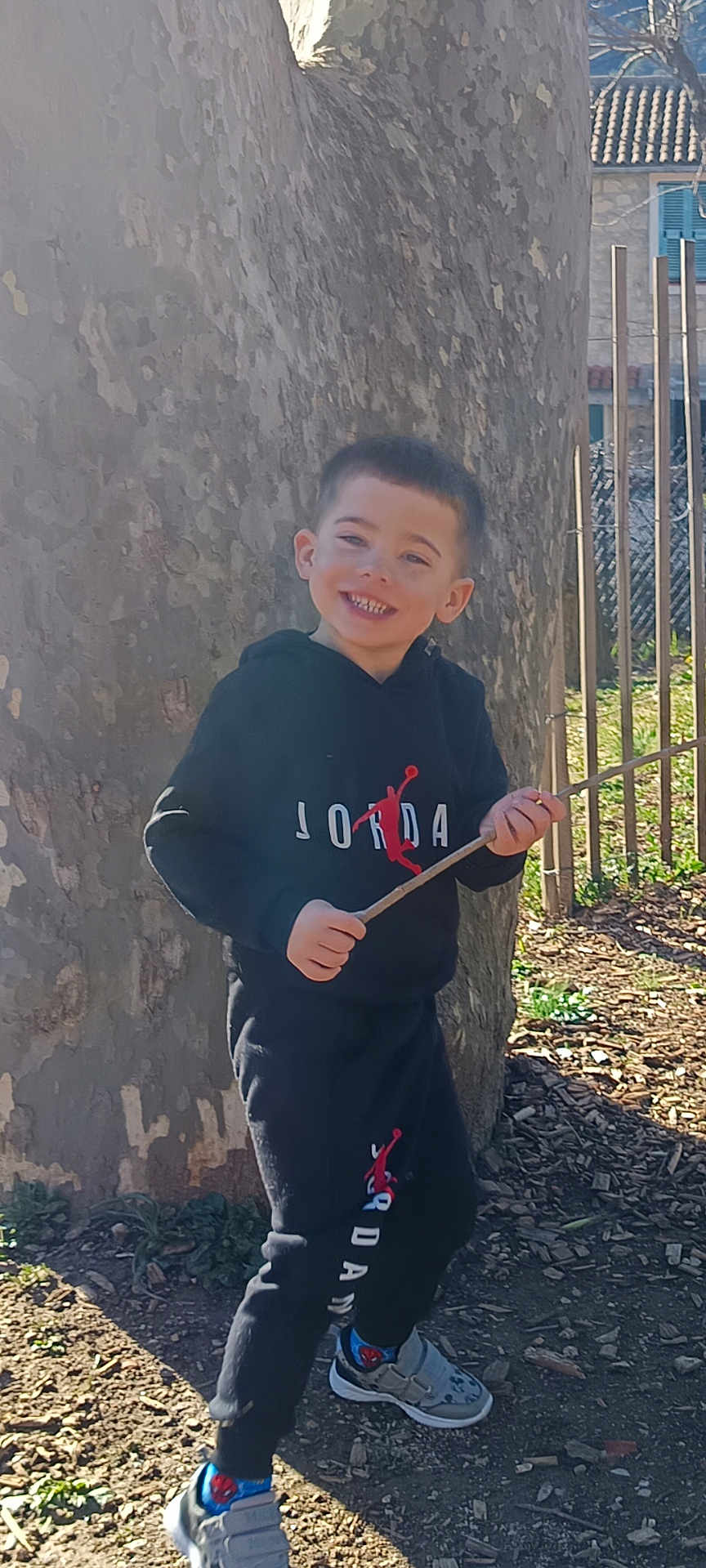Benjamin a rejoint le concours — aidez-le/la à gagner de superbes lots ! child, boy, smiling, tree_trunk, stick, outdoors, hoodie, sweatpants, sneakers, park, fence, sunlight, happy, portrait, standing, play, ground_cover, leaf_litter, casual_clothing, logo