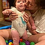 baby, child, woman, person, ball_pit, colorful_balls, dinosaur_onesie, indoor, bookshelf, sitting, smiling, holding, legs, face, playtime, toys, happy, family, cute, fun