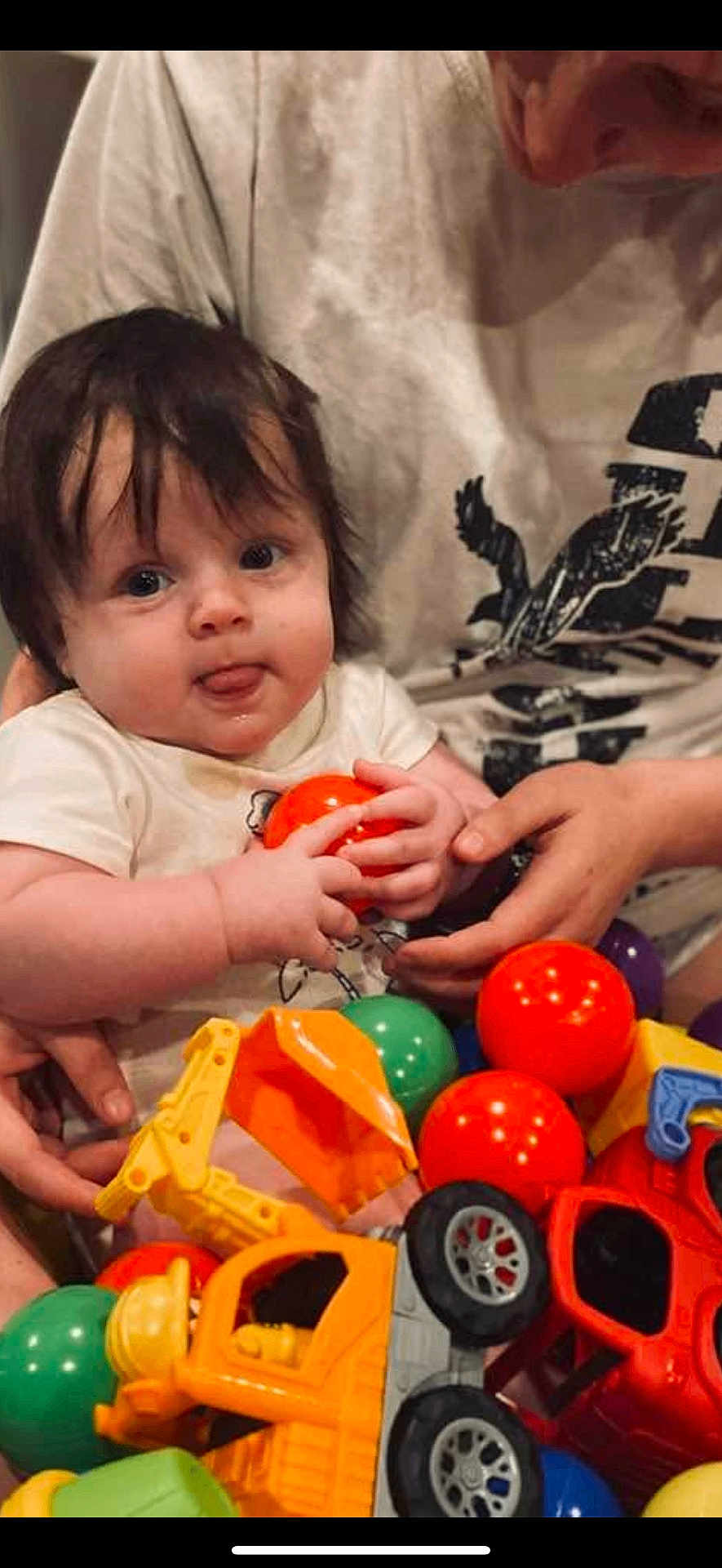 Asher joined the competition — help win amazing prizes! baby, child, toy, orange_ball, green_ball, red_toy_car, yellow_toy_excavator, person, hands, shirt, face, tongue_out, indoor, playtime, cute, smiling, holding, colorful, plastic_toys, casual_clothing