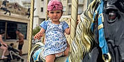 Valentina participe au concours pour gagner de l'argent avec cette photo : toddler, child, carousel, horse, ride, pink_cap, blue_dress, sandals, urban, background, people, building, pole, outdoor, playground, serious_face, toy, amusement_ride, summer, fun