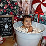 baby, marshmallows, bucket, christmas_tree, holiday_decor, peppermint_lollipop, sign, hot_chocolate, table, wood_background, red_ribbon, festive, smiling, indoors, cute, child, face, winter, drink, celebration