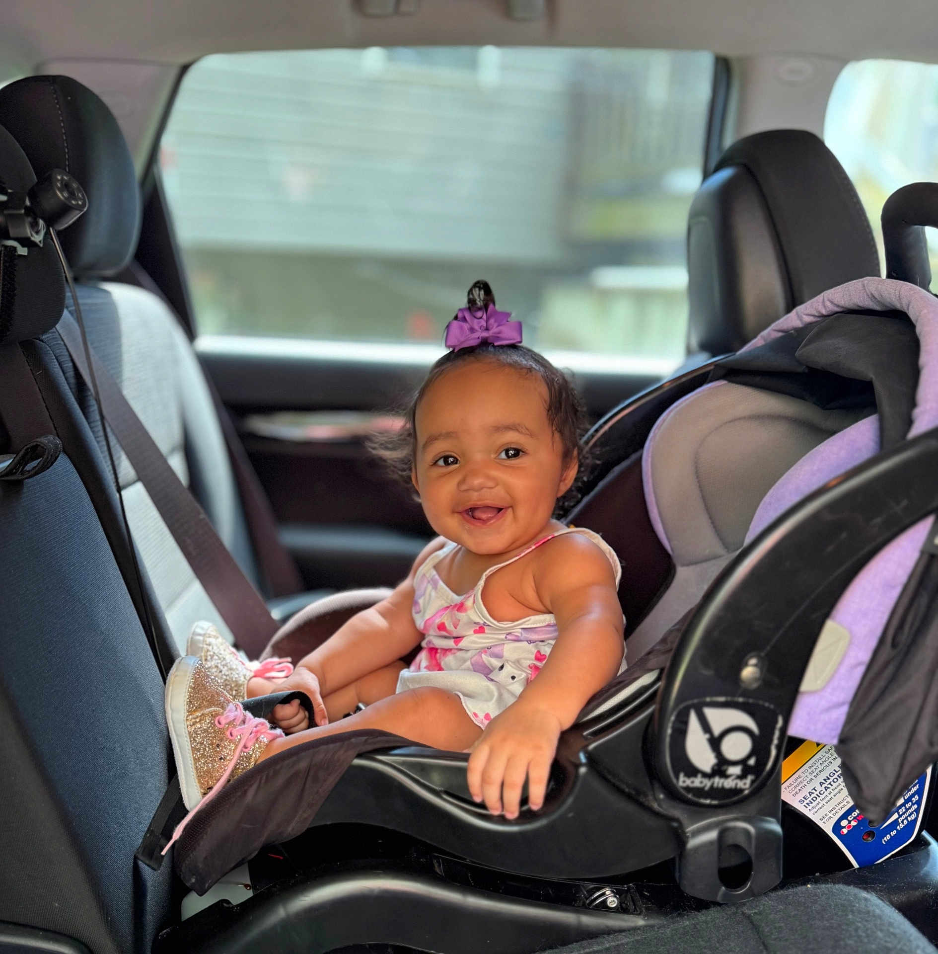 Elena is registered to the contest to win money with this photo: baby, car_seat, child, smile, happy, indoor, seat, vehicle, clothing, footwear, purple_bow, infant, child_safety, car_interior, seatbelt, person, cute, young_child, portrait, babytrend