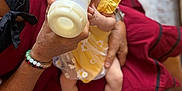 Mélodie participe au concours pour gagner de l'argent avec cette photo : baby, bottle, feeding, yellow_dress, adult_hand, arm, person, infant, milk, clothing, sitting, looking, curious, skin, fingers, floor, footwear, bracelet, fabric, indoors