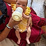 baby, bottle, feeding, yellow_dress, adult_hand, arm, person, infant, milk, clothing, sitting, looking, curious, skin, fingers, floor, footwear, bracelet, fabric, indoors