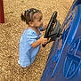 toddler, child, playground, steering_wheel, blue_play_structure, wood_chips, grass, outdoor, curly_hair, hair_ties, playing, side_view, casual_clothing, short_sleeves, imaginative_play, young_child, daylight, park, fun, active