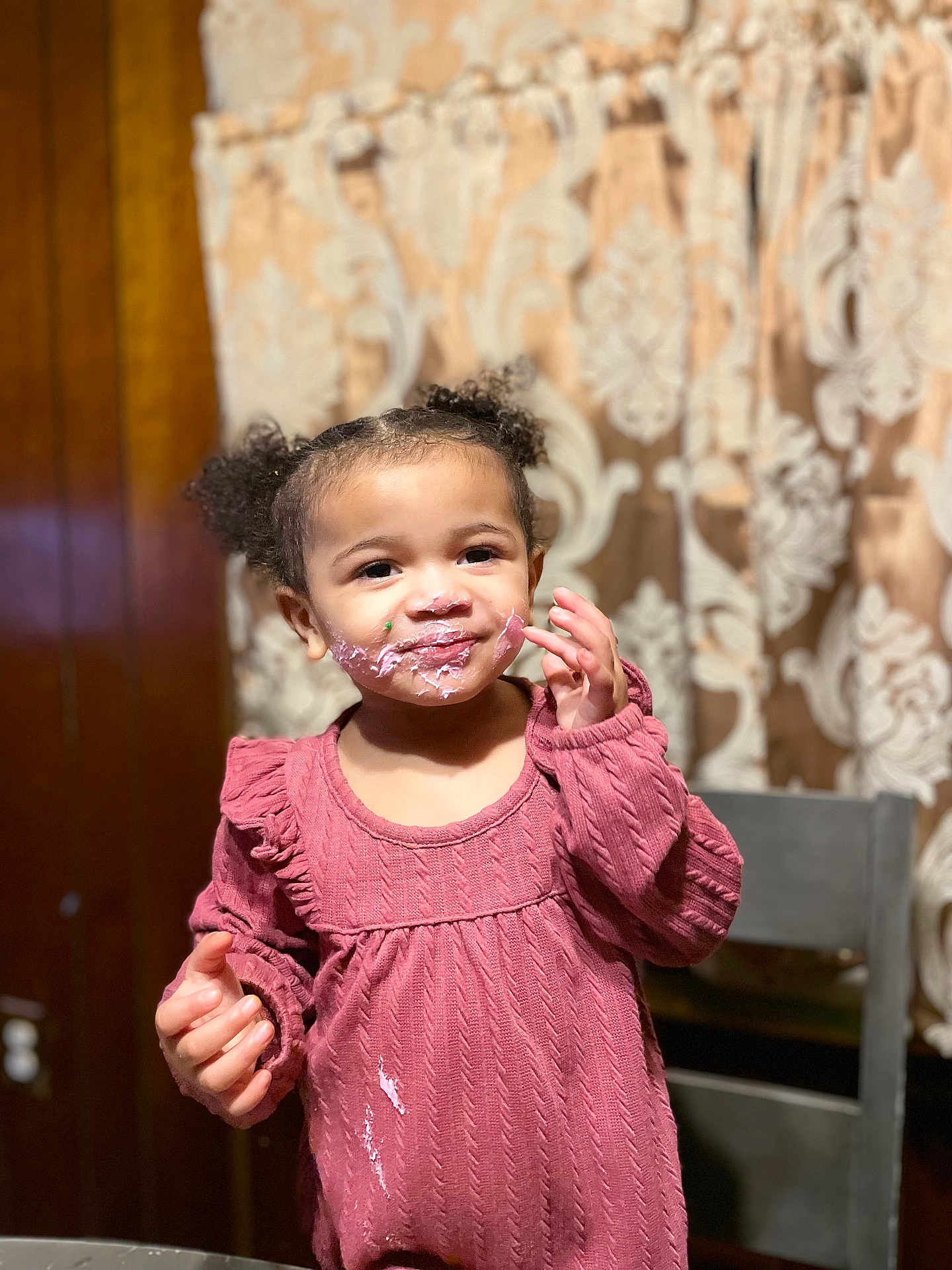 Zylah Brown joined the competition — help win amazing prizes! toddler, child, indoor, pink_frosting, messy_face, pink_sweater, curly_hair, patterned_curtain, wooden_chair, smile, face, person, cute, young_child, portrait, expression, clothing, frosting, hand, happy